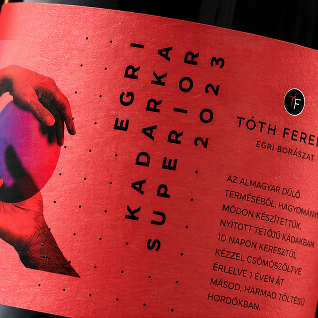 Close-up of a red wine label with black text reading 'EGRI KADARKA SUPERIOR 2023' and 'TÓTH FERÉC', plus a hand holding a blue-purple fruit illustration on the left.