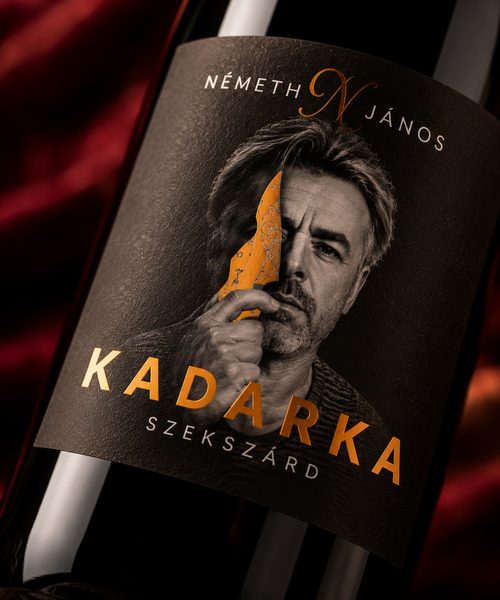 A close-up of a wine bottle label featuring a portrait of a man holding a knife, with the text 'NÉMETH JÁNOS' at the top and 'KADARKA SZEKSZÁRD' in large gold lettering at the bottom. The background is dark with red draped fabric.